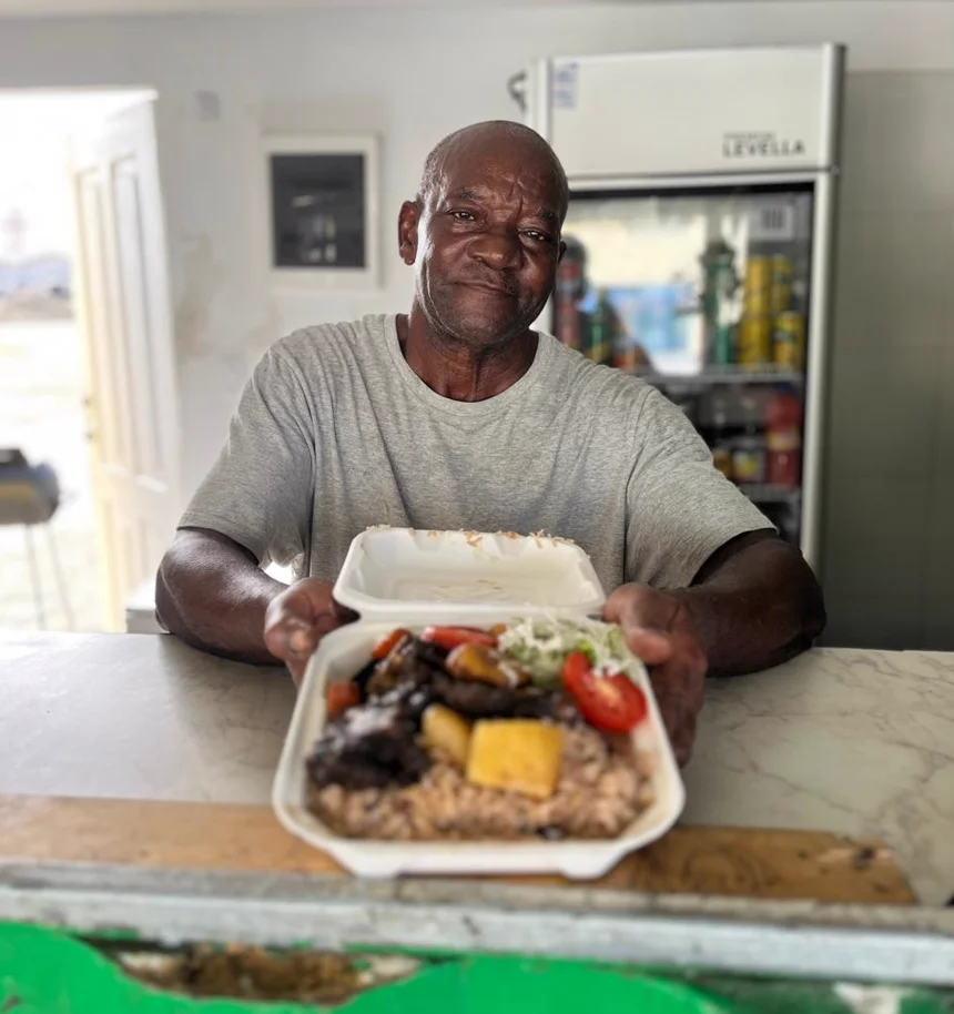 The Jamaican cook at Reggae Vibes Takeaway holding a takeaway box of oxtail with rice and peas, fried plantain and salad
